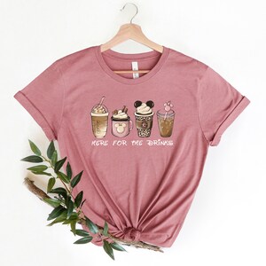 May include: A mauve t-shirt with a graphic of five different coffee drinks, including a Mickey Mouse-shaped cup. The text "Here for the drinks" is printed below the graphic.