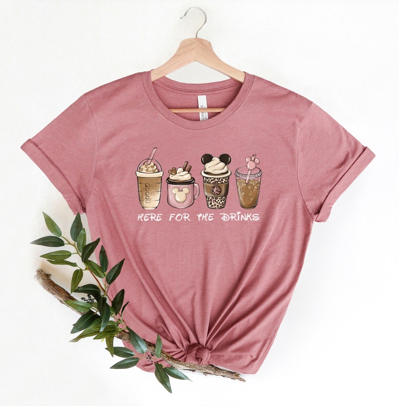 May include: A mauve t-shirt with a graphic of five different coffee drinks, including a Mickey Mouse-shaped cup. The text "Here for the drinks" is printed below the graphic.