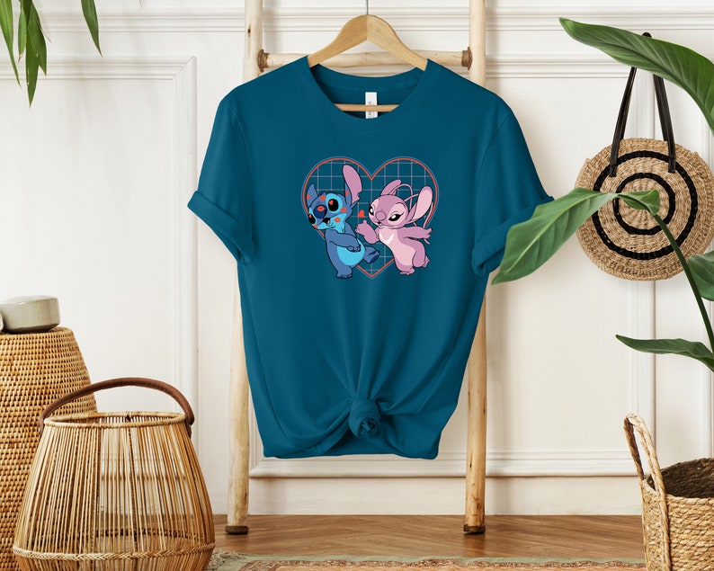 Puede incluir: Camiseta azul turquesa con un gr&aacute;fico de Stitch y Angel de Lilo & Stitch dentro de un coraz&oacute;n. El coraz&oacute;n tiene un patr&oacute;n de cuadr&iacute;cula y corazones rojos.
