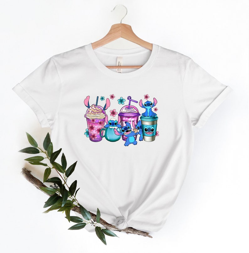 May include: White t-shirt with a graphic of blue cartoon characters holding colorful cups of coffee and iced coffee. The characters are surrounded by pink flowers.