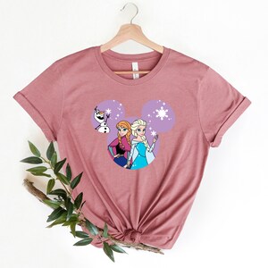 May include: A mauve t-shirt with a purple Mickey Mouse head design featuring the characters Elsa and Anna from Frozen and Olaf. The design includes snowflakes and sparkles.