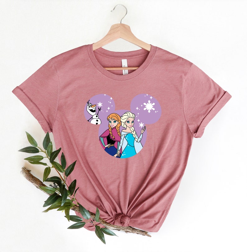 May include: A mauve t-shirt with a purple Mickey Mouse head design featuring the characters Elsa and Anna from Frozen and Olaf. The design includes snowflakes and sparkles.