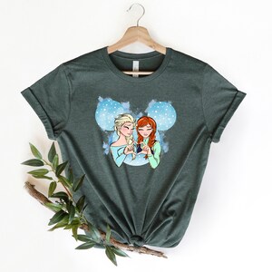 May include: A dark green t-shirt with a graphic of Elsa and Anna from Frozen making a heart shape with their hands. The graphic is set against a blue and white background with snowflakes.