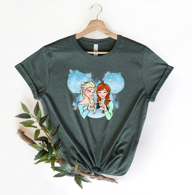 May include: A dark green t-shirt with a graphic of Elsa and Anna from Frozen making a heart shape with their hands. The graphic is set against a blue and white background with snowflakes.