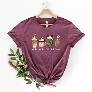 May include: A maroon t-shirt with a graphic of four different coffee drinks. The text "Here for the drinks" is printed below the drinks.