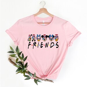 May include: Pink t-shirt with a graphic of Mickey Mouse, Minnie Mouse, Donald Duck, Goofy, and Pluto wearing red, white, and blue patriotic hats and sunglasses. The text "FRIENDS" is below the graphic.