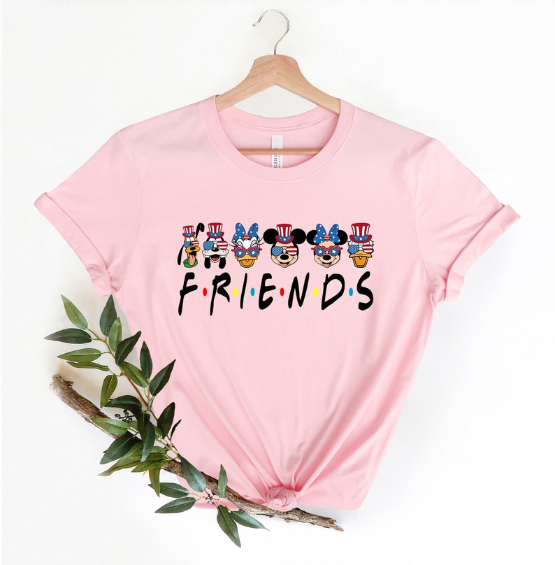 May include: Pink t-shirt with a graphic of Mickey Mouse, Minnie Mouse, Donald Duck, Goofy, and Pluto wearing red, white, and blue patriotic hats and sunglasses. The text "FRIENDS" is below the graphic.