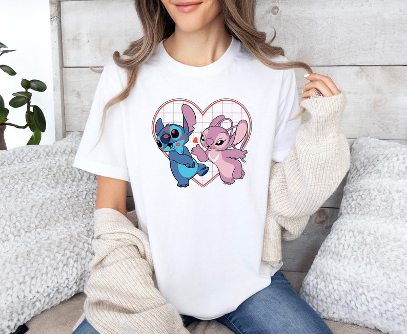 Puede incluir: Camiseta blanca con un gr&aacute;fico de Stitch y Angel de Lilo & Stitch. Los personajes est&aacute;n dentro de un coraz&oacute;n rojo con un patr&oacute;n de cuadr&iacute;cula.