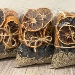 Könnte beinhalten: Drei durchsichtige Plastikbeutel gefüllt mit getrockneten Orangenscheiben, dunklen Beeren und Lavendelblüten. Jeder Beutel ist in der Mitte mit einer braunen Juteschnur zu einer Schleife gebunden, die den Inhalt mit Früchten oben und Lavendel unten sichert.