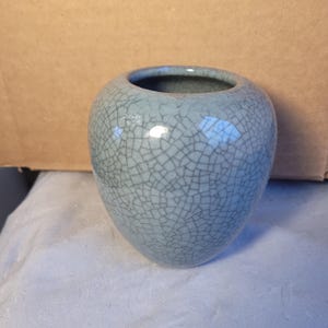 Może przedstawiać: Mały, jasnoniebieski ceramiczny wazon z wykończeniem w postaci spękań. Wazon ma zaokrąglony korpus i szeroki otwór. Powierzchnia pokryta jest siecią cienkich, ciemnych linii, tworząc unikalną fakturę. Wazon jest prawdopodobnie przeznaczony do celów dekoracyjnych.