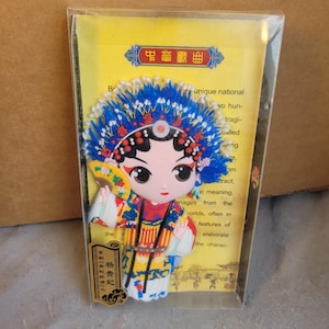 Peut inclure: Un aimant coloré représentant une figurine de style cartoon en costume d'opéra chinois traditionnel. La figurine porte une coiffe bleue et tient un éventail. Le fond contient du texte en chinois et en anglais.