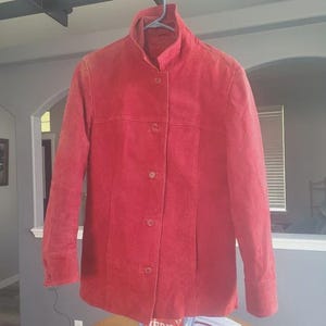 May include: A red suede jacket with a mandarin collar and button-down front. The jacket has long sleeves with buttoned cuffs. The garment is hanging on a hanger.