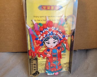 May include: A colorful Beijing opera figure magnet, encased in clear plastic. The figure is dressed in a red and blue costume with elaborate headwear. The packaging includes text about Beijing opera.