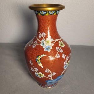 May include: A small, decorative vase with a reddish-orange background and floral designs. The vase has a gold-colored rim and base. The floral design includes white, blue, and pink flowers with green leaves. The vase is likely made of ceramic or porcelain.