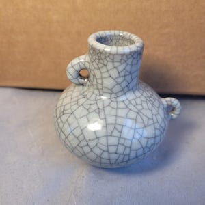 Może przedstawiać: Mały, kremowobiały ceramiczny wazon z wzorem spękanego szkliwa. Wazon ma zaokrąglony korpus, cylindryczną szyję i dwa małe uchwyty w kształcie pętli. Efekt spękania tworzy sieć cienkich linii na powierzchni.
