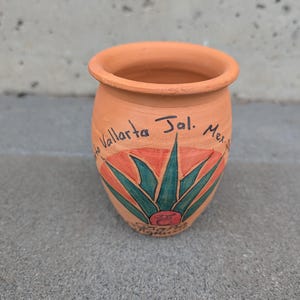 Op de afbeelding: Een kleine, terracotta-kleurige keramische pot met een handgeschilderd agaveplantontwerp in groen, oranje en rood. De tekst "Puerto Vallarta Jal. Mex." staat rond de pot geschreven. De pot heeft een afgeronde body en een wijde opening.