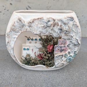 May include: A white ceramic decorative bowl with a textured exterior and a cut-out revealing a miniature diorama. The diorama features a small white building with blue accents, surrounded by dried flowers and a pink and red flower.