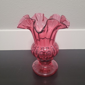 Puede incluir: Un jarrón de cristal rosa con un borde ondulado. El jarrón tiene un cuerpo bulboso y una base redonda. El cristal tiene un acabado brillante y es de un tono rosa intenso. El jarrón es probablemente vintage y adecuado para exhibir flores.