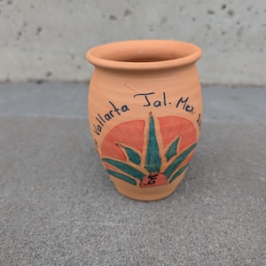 May include: A small, terracotta-coloured ceramic pot with a hand-painted design. The design features a red and green agave plant with the text "Vallarta Jal. Mex." written in black ink. The pot has a rounded body and a slightly flared rim.
