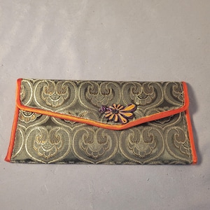 May include: An olive green clutch with a gold floral pattern and orange trim. The flap closure features a decorative flower with purple and yellow accents. A stylish accessory for carrying essentials.
