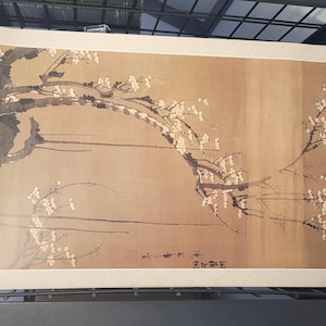 Pode incluir: Uma pintura de estilo japonês com um galho de flor de ameixa com flores brancas e galhos cinza escuro em um fundo bege. A obra de arte inclui caracteres japoneses no canto inferior direito.