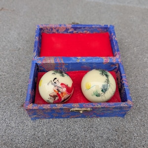 Puede incluir: Un par de bolas chinas de salud de color crema con escenas pintadas, presentadas en una caja decorativa azul y roja. Una bola representa una figura tocando un instrumento, la otra un paisaje. La caja está abierta.