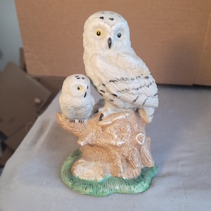 May include: A ceramic owl figurine featuring a large owl perched on a tree stump with a smaller owl beside it. The owls are primarily white with black markings and yellow eyes. The tree stump is brown with green grass at the base.