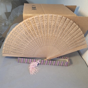 May include: A light brown, intricately carved wooden hand fan, partially open, with a matching box featuring colorful stripes and a pink tassel. The fan's design includes delicate cut-out patterns. The fan is a decorative accessory.
