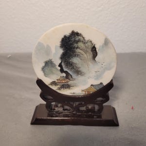 May include: A round, off-white decorative plate with a landscape painting of mountains, trees, and buildings. The plate is displayed on a dark brown wooden stand with a rectangular base. The painting features shades of blue, green, and brown.