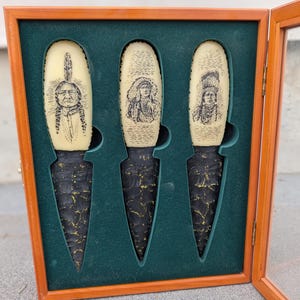 May include: Set of three decorative knives in a wooden display case. The knives have cream-colored handles with engraved Native American portraits and black, textured blades. The case has a green velvet interior.