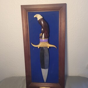 May include: Framed decorative dagger featuring a carved eagle perched atop the handle. The handle is dark brown with gold accents and a purple band. The blade is silver, and the frame is dark wood, set against a blue background.