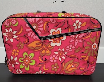Puede incluir: Una maleta rectangular rosa con estampado floral, ribetes negros y cremallera negra. La maleta presenta un diseño floral vibrante en tonos rosa, naranja, verde y blanco. El texto "Loads of Fun" está arriba.