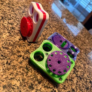 Könnte beinhalten: Drei bunte Zappelspielzeuge. Eines ist weiß mit roten Streifen und einem roten Rad. Die anderen beiden sind grün und lila mit kreisförmigen Designs. Die Spielzeuge liegen auf einer gesprenkelten braunen Oberfläche.