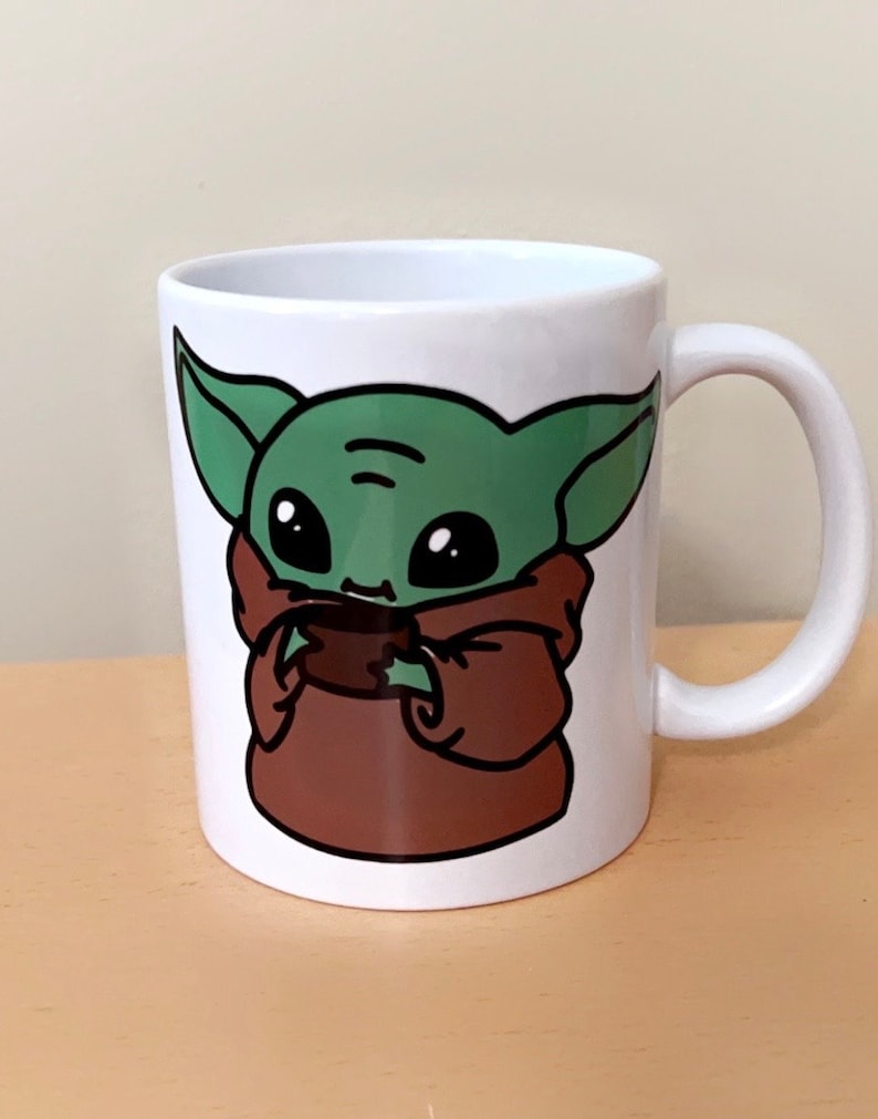 May include: White ceramic mug featuring a cartoon illustration of a green alien character with large black eyes and brown robes. The character is holding a small brown object. The mug has a white handle.
