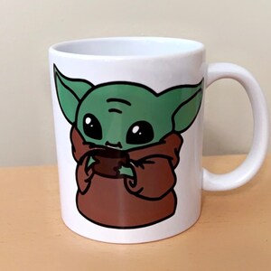 May include: White ceramic mug featuring a cartoon illustration of a green alien character with large black eyes and brown robes. The character is holding a small brown object. The mug has a white handle.