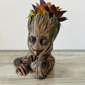 May include: A brown Groot-shaped planter with a smiling face, holding its chin. The planter is filled with a succulent plant with dark red and yellow leaves. The planter is made of a textured material.