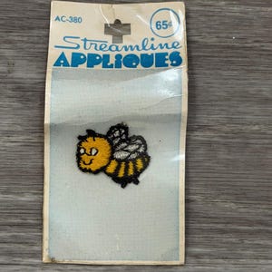 May include: A vintage Streamline Appliques patch featuring a smiling cartoon bee. The bee is yellow, black, and white, with the text "Streamline APPLIQUES" in blue. The patch is in its original packaging.