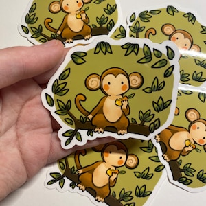 May include: A collection of stickers featuring a cartoon monkey. The monkey is brown with a tan face and ears, sitting on a brown branch and holding a yellow fruit. The background is a light green with dark green leaves.