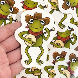 Könnte beinhalten: Eine Sammlung von Aufklebern mit einer Cartoon-Frösche, die Banjo spielen. Der Frosch ist grün, trägt einen braunen Cowboyhut und ein rotes Halstuch. Das Banjo ist beige und braun. Die Aufkleber befinden sich auf weißem Hintergrund.