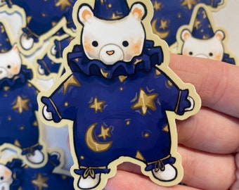 Puede incluir: Una pegatina con un oso blanco vestido con un atuendo azul oscuro con estrellas doradas y lunas crecientes. El oso lleva un sombrero a juego y un cuello con volantes. La pegatina tiene un borde color crema.
