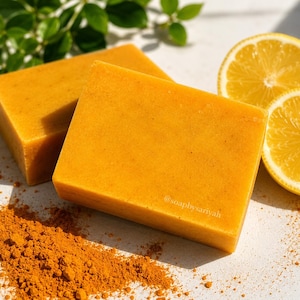 May include: Two square, bright orange bars of soap are displayed with two lemon halves and a pile of orange powder. The soap has a smooth texture. Green leaves are in the background. The soap is labeled with the text "@soapsbysariyah".