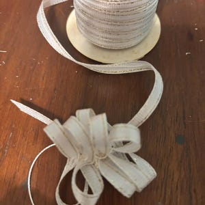 May include: A spool of white ribbon with gold trim, partially unraveled, with a small bow tied at the end. The ribbon is on a wooden surface. The ribbon is likely used for gift wrapping or crafting.