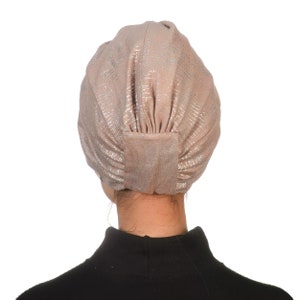 May include: A light brown turban with silver shimmer, worn on a person's head. The turban is made of a soft, stretchy fabric.