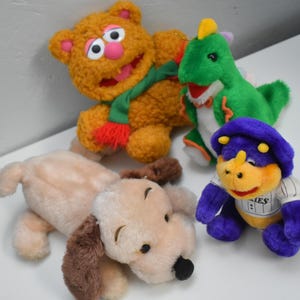 May include: A collection of plush toys, including a brown bear with a green scarf, a green dragon, a purple and yellow dinosaur, and a tan dog with brown ears. The toys are soft and colorful.