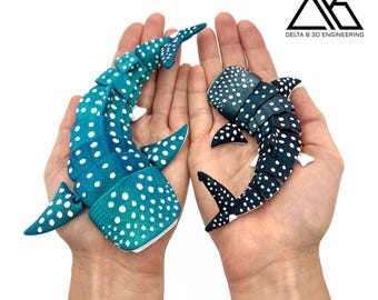 May include: Two articulated whale shark toys, one teal and one navy, each with white polka dots. The toys are held in open hands against a white background. The logo "DELTA B 3D ENGINEERING" is in the upper right corner.