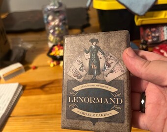 May include: A brown box with a vintage design featuring a man in a blue suit holding a deck of cards. The box says "Lenormand Oracle Cards" and "Alexandre Musruck".