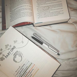 May include: An open book with text, a notebook with the words "PART ONE" and a list of terms, and two pens. The notebook has a dragon illustration. The scene is on a white, textured surface.