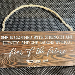 May include: A wooden sign with a brown woodgrain finish and white text. The sign reads "She is clothed with strength and dignity, and she laughs without fear of the future. Proverbs 31:25".