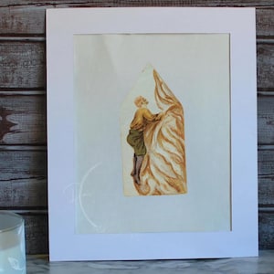 May include: Watercolor painting of a figure climbing a textured, golden-brown structure. The figure is dressed in shades of yellow and green. The artwork is framed in a white mat, set against a wooden background.