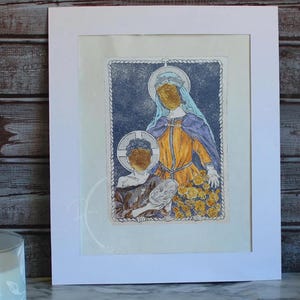 May include: An art print featuring a religious image of two figures with golden halos. The figures are depicted in a stylized manner, with one wearing a yellow and blue robe and the other holding a mask. The print is framed in a white mat.